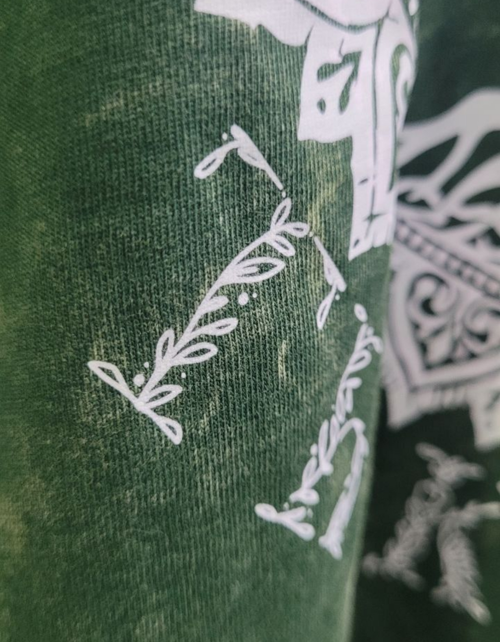 Close-up of macro detailing of a dark green acid-wash t-shirt with a large white graphic on the back featuring a tree of life, The KalpVriksh, and the words 'Manifest Abundance The KalpVriksh'.